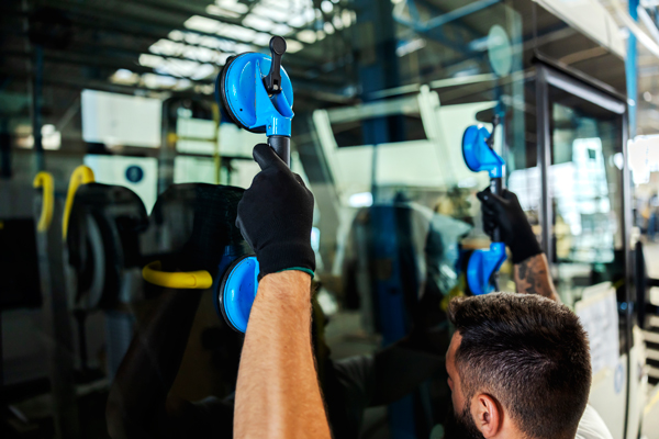 A technician from British Bus Repair Services (BBRS) is installing or repairing a glass panel on a bus using blue suction cup lifters. The worker, wearing black protective gloves, is carefully positioning the glass in place inside a well-lit workshop. The bus’s interior and exterior are visible in the background, highlighting the professional repair environment.